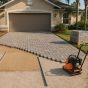 Paver driveway being installed in Jacksonville: compacted base, screeded bedding sand between rails and concrete pavers laid from the garage, with a plate compactor nearby