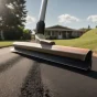 a homeowner smooths fresh asphalt on a driveway with a large push squeegee, under a sunny sky.